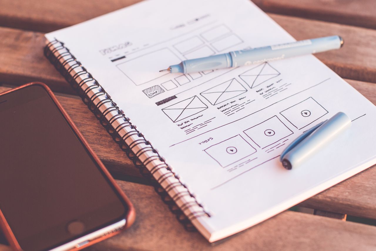 services-02 Close-up of a notebook with wireframe sketches and a smartphone on a wooden desk.